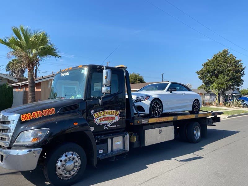 Expedite Towing flatbed tow truck with a red car safely secured on its bed. The truck is parked on a deserted road with the San Diego skyline visible in the distance.